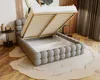 A dark gray upholstered bed frame with the mattress base lifted, revealing storage space underneath. The bed is placed on a rug in a room with wooden floors and curtains. Náhled obrázku produktu