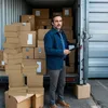 A man stands in front of an open shipping container filled with boxes, holding a clipboard. Voir les images du produit