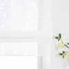 A close-up view of a white Roman blind with a subtle branch pattern, partially drawn, against a white wall. A small branch with light yellow flowers and green leaves is placed to the right of the blind. Produktbild-Vorschau