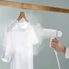 A hand is holding a white garment steamer and steaming a white t-shirt hanging on a wooden hanger. Produktbild-Vorschau