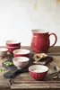 A rustic still life featuring red ceramic bowls and a pitcher filled with a creamy substance, accompanied by bread slices and greenery, arranged on a wooden surface. Produktbild-Vorschau
