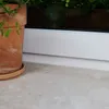 A close-up view of a terracotta pot with a plant on a marble-like surface next to a white baseboard and window frame. Produktbild-Vorschau