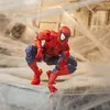 A Spider-Man action figure is posed in a crouching position on a surface covered in fake spiderwebs, with a blurred background suggesting a building. Podgląd zdjęcia produktu
