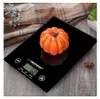 A black digital kitchen scale with a small orange pumpkin on top, placed on a wooden surface with a folded blanket and some dried flowers in the background. Náhľad obrázku produktu