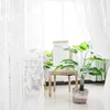 A white lace curtain is draped in the foreground, with a chair and a potted plant in the midground. A paper bag with text is leaning against the chair. Produktbild-Vorschau