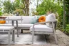 A modern outdoor lounge set with a sofa, armchair, and coffee table on a patio with a rug and lush greenery in the background. Produktbild-Vorschau