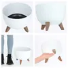 A collage of four images showing a white planter pot with wooden legs from different angles. One image shows the inside of the pot, another shows the pot from the side, a third shows the pot with a person's legs next to it for scale, and the fourth shows t Náhled obrázku produktu
