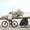 A man stands next to a bicycle with a child trailer attached, outdoors. Produktbild-Vorschau
