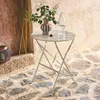 A round, beige bistro table with a vase of greenery, a glass bottle, and a glass on it, set on a stone patio next to a rustic wooden door and a woven lantern. Produktbild-Vorschau