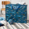 A large storage bag with a shark pattern is placed on a rug next to a chair. Some folded towels and a blue knitted item are on the floor in front of the bag. A blue ball with shark patterns is also on the floor. Produktbild-Vorschau