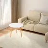 A round white coffee table with wooden legs is placed on a cream rug in front of a beige sofa with a green throw blanket and a light gray pillow. Produktbild-Vorschau