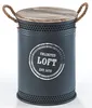 A dark gray cylindrical storage bin with a wooden lid, rope handles, and perforated detailing at the top and bottom. It has a circular graphic with text on the front. Produktbild-Vorschau