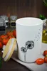 A white ceramic storage jar with a dandelion design and a bamboo lid, placed on a white cloth with tomatoes and dill in the background. Produktbild-Vorschau