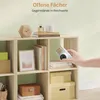 A hand is placing a white digital camera onto a shelf of a light oak cube storage unit. The shelves contain books, a framed picture, and decorative items. Produktbild-Vorschau