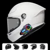A white motorcycle helmet with a Bluetooth headset attached, displayed against a black background. Several other motorcycle helmets are shown in a row at the bottom. Podgląd zdjęcia produktu