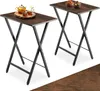 Two folding side tables with dark wood-effect tops and black metal frames, set against a white background. One table has a teapot and two small cups on it, while the other has two bowls of snacks. Náhled obrázku produktu