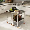 A stainless steel work table with two shelves and wheels, set in a modern kitchen environment. The table is topped with a blender, a bowl of fruit, and a plate of green produce. The lower shelves hold baskets filled with various fruits and vegetables. Produktbild-Vorschau