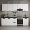 A modern, white kitchen unit with a black countertop and appliances against a gray concrete wall. Produktbild-Vorschau