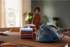 A steam generator iron is placed on a wooden table next to a stack of folded shirts. A woman is visible in the background. Produktbild-Vorschau