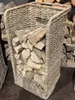 A wicker basket filled with firewood, placed on a brown carpet with stacks of firewood in the background. Produktbild-Vorschau