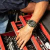 A person's hand with a green digital watch is reaching into a red toolbox filled with tools. Produktbild-Vorschau