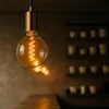 A close-up of a vintage-style filament light bulb hanging in what appears to be a cafe or restaurant setting, with shelves of mugs visible in the background. Produktbild-Vorschau