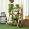 A wooden plant stand with various potted plants on it, placed on a green lawn in front of a white wooden fence. Produktbild-Vorschau