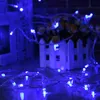 Blue string lights are draped over a wooden surface in front of a white picket fence and some flowers. Náhľad obrázku produktu