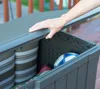 A hand is closing the lid of a brown plastic storage box on a wooden deck. Inside the box are cushions and a soccer ball. Produktbild-Vorschau