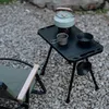 A camping table with a kettle and cups set up on a gravel surface next to a camping chair. Produktbild-Vorschau