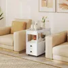 A white rolling side table with two drawers and open shelves is placed next to a beige armchair in a living room setting. The table is decorated with a teapot, cups, flowers, and books. Produktbild-Vorschau