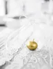 A golden apple candle inside a clear glass orb with a white ribbon, resting on a white lace tablecloth. Podgląd zdjęcia produktu