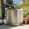 A person is holding the lid of a Solo Stove fire pit, with logs nearby. Produktbild-Vorschau