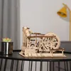A wooden marble run model is displayed on a black table, with a small potted plant to the left and a yellow lamp in the blurred background. Produktbild-Vorschau