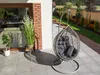 A hanging egg chair with cushions sits on a patio next to a house, with plants and a garden in the background. Náhled obrázku produktu
