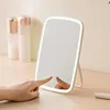A hand is touching a makeup mirror with LED lights on a wooden desk, with cosmetic bottles in the background. Podgląd zdjęcia produktu