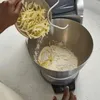 A person is adding shredded cheese to a stand mixer bowl filled with flour. Produktbild-Vorschau