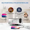 A digital alarm clock with a projection feature, shown with various functions illustrated around it, including phone charging, temperature and humidity display, and mirror usage. Náhled obrázku produktu