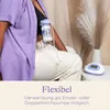A person is using a breast pump while wearing a purple shirt and cream-colored pants. A breast pump and collection bottle are attached to the person's chest, with tubing leading to an electric breast pump unit on a table. A decorative dried plant is in the Produktbild-Vorschau
