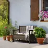 A garden bench with storage is placed on a patio next to potted plants and a window with flowers. Voir les images du produit