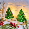 Two ceramic Christmas tree night lights plugged into an outlet, with festive decorations in the foreground. Náhľad obrázku produktu