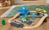 A toy train set with a gas station is set up on a rug and wooden floor, with gift-wrapped boxes in the background. Produktbild-Vorschau