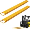 Two yellow forklift extensions are shown in the foreground, with a yellow and black forklift in the background. Náhľad obrázku produktu