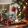 A Christmas-themed still life with a gift box, ornaments, a pinecone, and a lit candle in a glass holder, with a decorated Christmas tree in the background. Produktbild-Vorschau