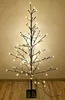 A decorative LED Christmas tree with a black trunk and branches, illuminated by warm white lights, standing on a wooden floor against a beige wall. Produktbild-Vorschau