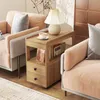 A wooden side table with drawers and shelves is placed between two pink armchairs, with a lamp and a cup of tea on top. Produktbild-Vorschau