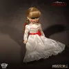 A doll dressed in a white dress with red accents, standing against a dark, textured background. Náhled obrázku produktu