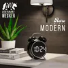 A black retro-style digital alarm clock is displayed on a white surface next to books and a potted plant, with a wall lamp and wood-paneled wall in the background. Náhled obrázku produktu