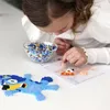A young girl is creating a perler bead design on a pegboard, with a bowl of beads and a finished blue parrot design nearby. Produktbild-Vorschau