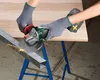 A person wearing gloves is using a cordless angle grinder to cut a metal profile. The metal profile is supported by a wooden board on a blue stand. Produktbild-Vorschau